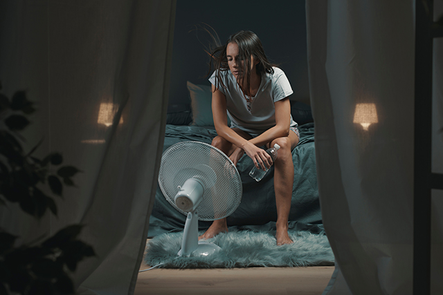 Exhausted woman suffering from hot flashes, she is holding a water bottle and sitting in front of a cooling fan in the bedroom