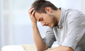 A young man sitting on a couch, leaning forward with his elbow on his knee and his hand on his forehead, appearing stressed or upset.