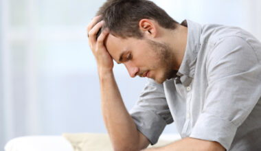 A young man sitting on a couch, leaning forward with his elbow on his knee and his hand on his forehead, appearing stressed or upset.
