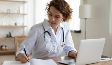 female doctor using laptop and writing notes in medical journal sitting at desk.