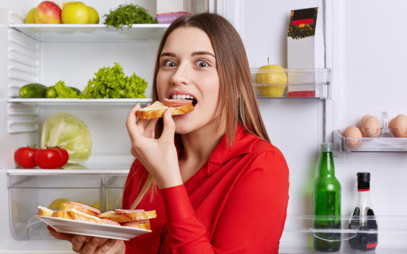 Young hungry woman eating in front of open fridge