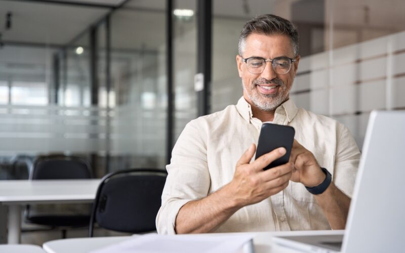 Middle aged man smiling at phone while sitting at office desk
