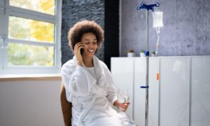 Woman sitting down on the phone with a glass of water while getting IV therapy