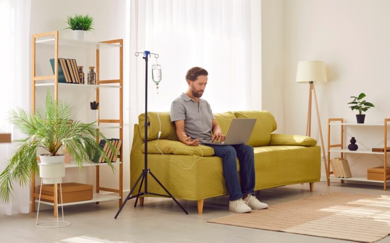 Man receiving IV therapy while sitting on couch with his laptop