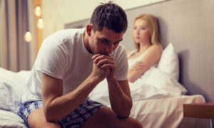 Man sitting on edge of bed upset while his girlfriend looks at him