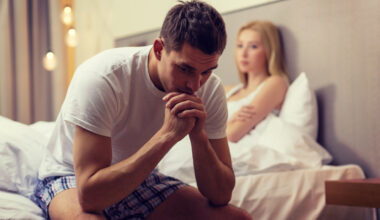 Man sitting on edge of bed upset while his girlfriend looks at him