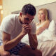 Man sitting on edge of bed upset while his girlfriend looks at him