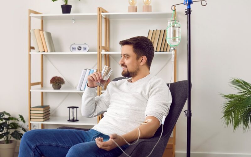 Man sitting down and receiving hydration IV therapy while drinking a glass of water