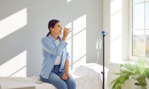 Woman with sterile tube in arm sitting by pole with IV bottle at medical center, drinking water and receiving vitamin therapy infusion to boost body immune system, preserve beauty or fight hangover