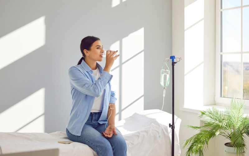 Woman with sterile tube in arm sitting by pole with IV bottle at medical center, drinking water and receiving vitamin therapy infusion to boost body immune system, preserve beauty or fight hangover