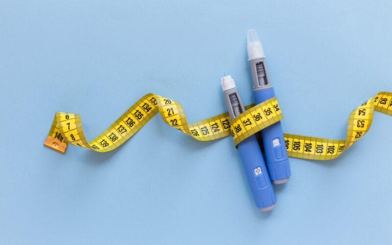 Two injectors dosing pens for subcutaneous injection of antidiabetic medication or anti-obesity medication hovering over a blue background. Yellow measuring tape around the injectors. antidiabetic