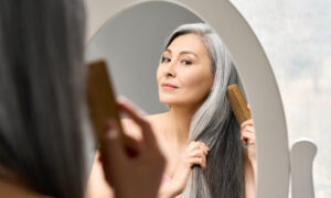 Senior attractive middle 50 years aged asian woman with gray hair looking at mirror reflection combing tangled gray hair. Alopecia hair loss prevention treatment after menopause advertising concept.
