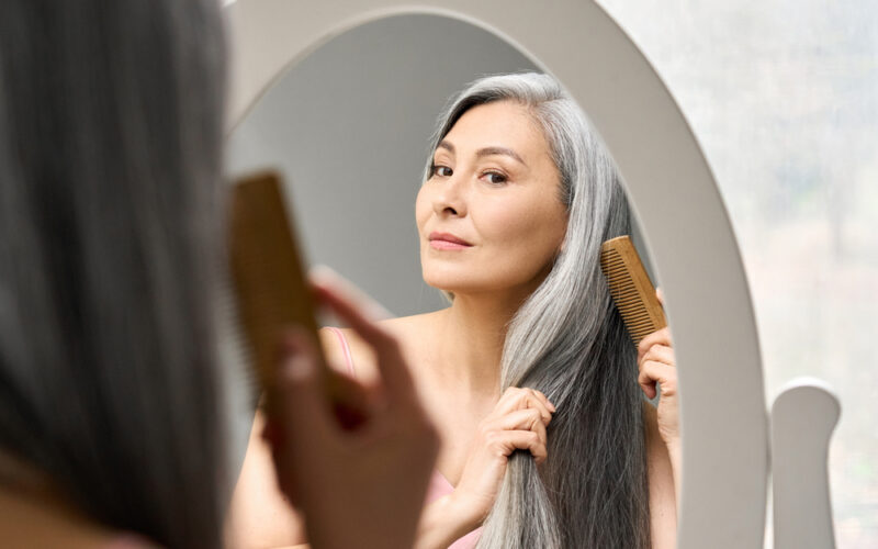 Senior attractive middle 50 years aged asian woman with gray hair looking at mirror reflection combing tangled gray hair. Alopecia hair loss prevention treatment after menopause advertising concept.