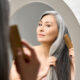 Senior attractive middle 50 years aged asian woman with gray hair looking at mirror reflection combing tangled gray hair. Alopecia hair loss prevention treatment after menopause advertising concept.
