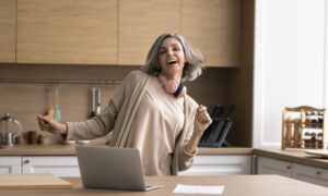 Happy mature woman enjoy track, dancing, stand at kitchen table with laptop, distracted from work, singing song, looking carefree alone at home. Modern tech, user of streaming digital music services