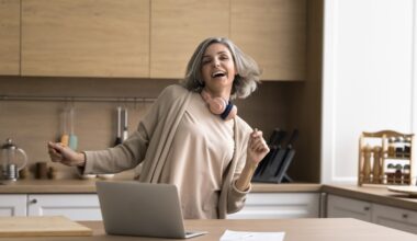 Happy mature woman enjoy track, dancing, stand at kitchen table with laptop, distracted from work, singing song, looking carefree alone at home. Modern tech, user of streaming digital music services