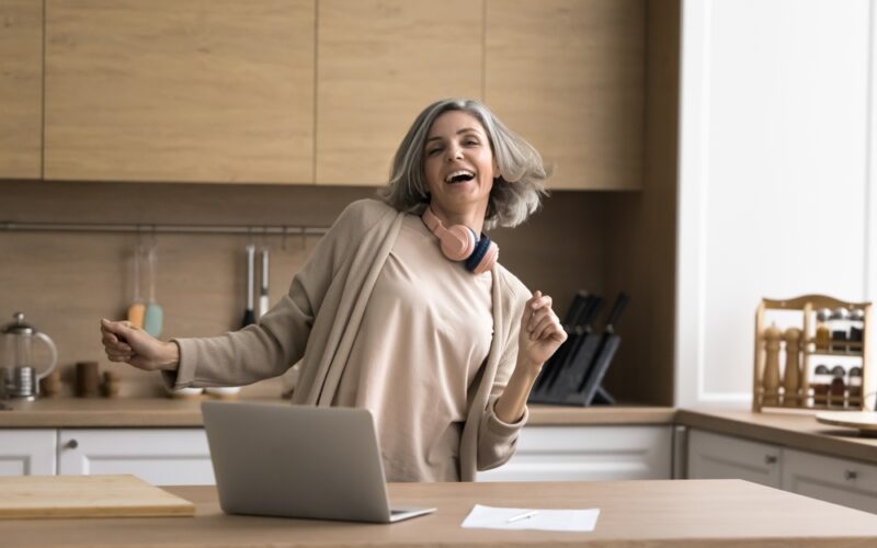 Happy mature woman enjoy track, dancing, stand at kitchen table with laptop, distracted from work, singing song, looking carefree alone at home. Modern tech, user of streaming digital music services