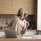 Happy mature woman enjoy track, dancing, stand at kitchen table with laptop, distracted from work, singing song, looking carefree alone at home. Modern tech, user of streaming digital music services