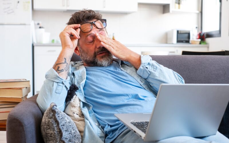 Tired businessman takes off glasses and feels eye strain from working online with laptop, Caucasian man sits on sofa and rubs his eyes, feels headache and lack of energy