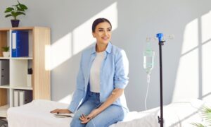 Young happy smiling woman sitting on the couch in medical clinic looking at the window while receiving IV drip infusion and vitamin injection therapy in her blood. Healthcare concept.