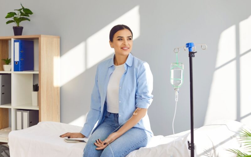 Young happy smiling woman sitting on the couch in medical clinic looking at the window while receiving IV drip infusion and vitamin injection therapy in her blood. Healthcare concept.