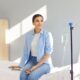 Young happy smiling woman sitting on the couch in medical clinic looking at the window while receiving IV drip infusion and vitamin injection therapy in her blood. Healthcare concept.