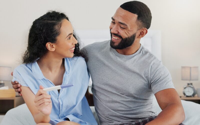 Happy couple, pregnancy stick and results in home, hug and support for positive baby news. People, plastic test and together for reading ovulation tool in bedroom, fertility treatment and embrace