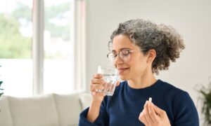 Happy healthy woman of middle age talking pill holding glass of water standing at home. Mature 50 years old lady taking vitamin health care supplement morning daily nutrition treatment concept