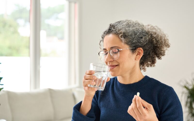 Happy healthy woman of middle age talking pill holding glass of water standing at home. Mature 50 years old lady taking vitamin health care supplement morning daily nutrition treatment concept