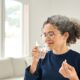 Happy healthy woman of middle age talking pill holding glass of water standing at home. Mature 50 years old lady taking vitamin health care supplement morning daily nutrition treatment concept