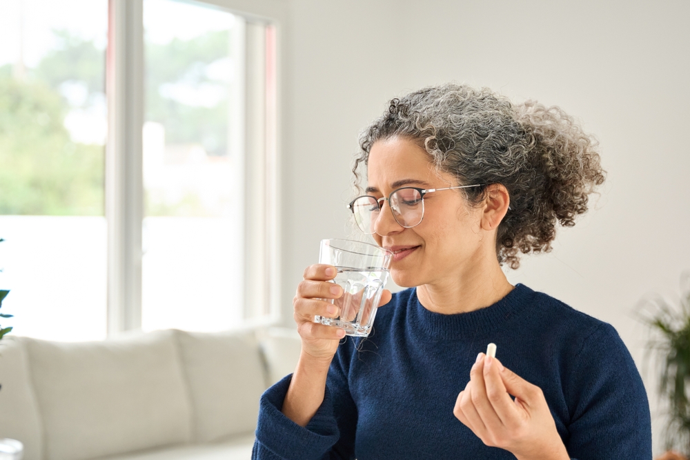 Happy healthy woman of middle age talking pill holding glass of water standing at home. Mature 50 years old lady taking vitamin health care supplement morning daily nutrition treatment concept