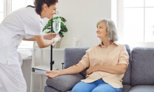 Senior woman with intravenous drip sitting on couch, physician, nurse doctor fixing IV drip infusion, geriatrics hospital eldercare, therapy to infuse vitamins, chemotherapy to maintain health clinic