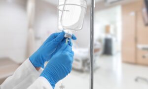 A close-up image of a healthcare professional wearing blue gloves adjusting an IV drip in a hospital setting, with the background showing a blurred patient bed and medical equipment.