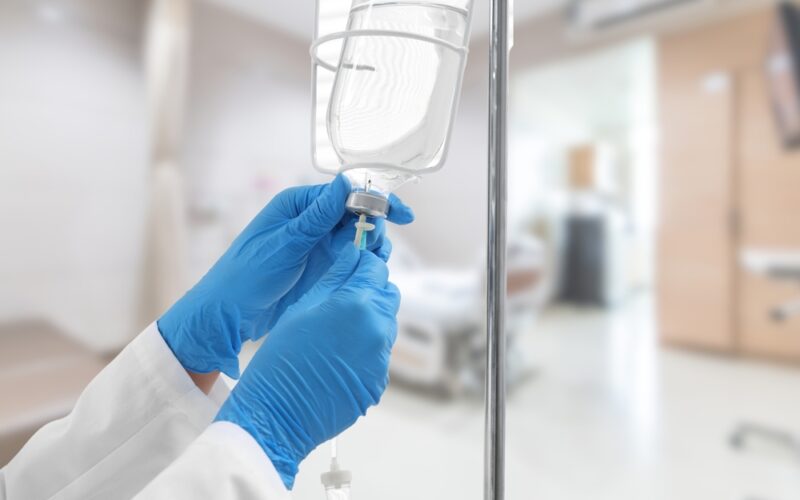 A close-up image of a healthcare professional wearing blue gloves adjusting an IV drip in a hospital setting, with the background showing a blurred patient bed and medical equipment.
