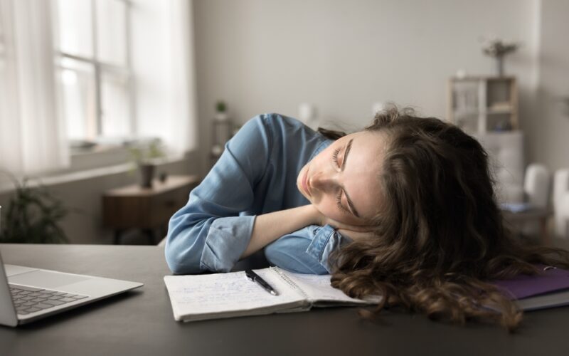 Drained young student woman sleeping, dozing lying down on desk, overloaded with studying, exams preparation, feels lack of energy after sleepless working night. Low productivity, overworking, fatigue