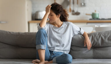 Exhausted frustrated young woman touching forehead, sitting on couch alone, suffering from strong headache or migraine, worried girl thinking about problems, divorce or break up, lost in thoughts