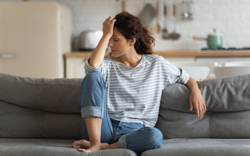 Exhausted frustrated young woman touching forehead, sitting on couch alone, suffering from strong headache or migraine, worried girl thinking about problems, divorce or break up, lost in thoughts
