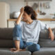 Exhausted frustrated young woman touching forehead, sitting on couch alone, suffering from strong headache or migraine, worried girl thinking about problems, divorce or break up, lost in thoughts