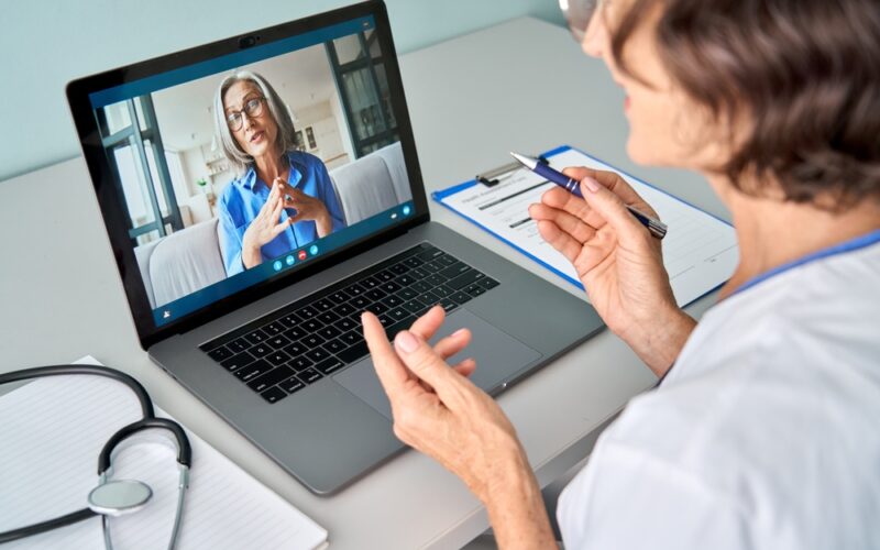 Female doctor therapist consulting older senior patient via virtual video call visit using laptop computer. Digital online healthcare, distance telemedicine. Telehealth videocall. Over shoulder view