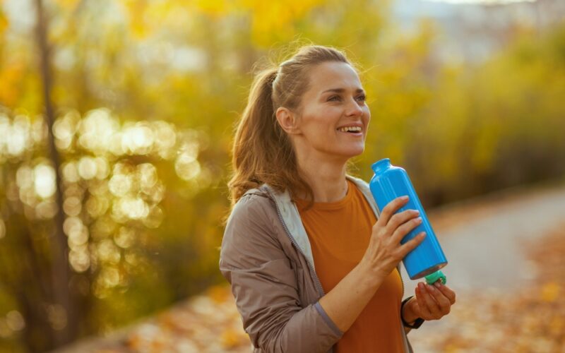 Hello autumn. happy trendy middle aged woman in fitness clothes in the park with bottle of water.