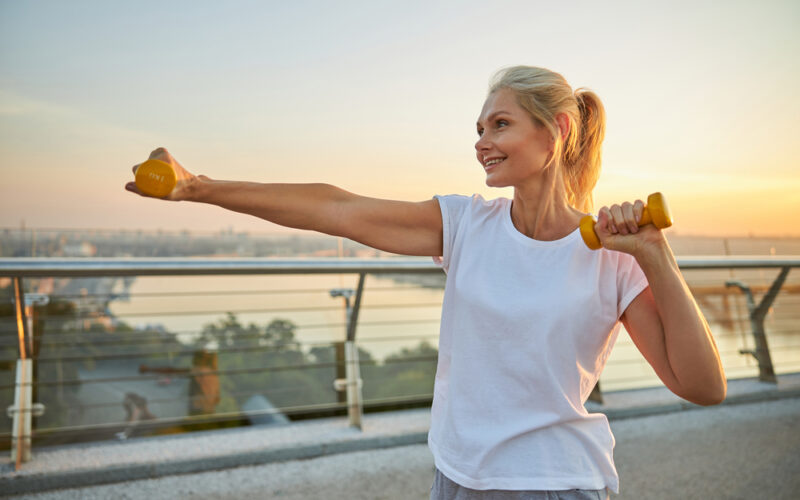 Smiling attractive energetic sporty blonde Caucasian female athlete boxing with dumbbells on a pedestrian bridge
