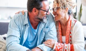 An adult couple in their late 40s, wearing glasses, smiling and embracing each other