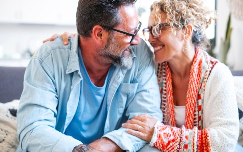 An adult couple in their late 40s, wearing glasses, smiling and embracing each other