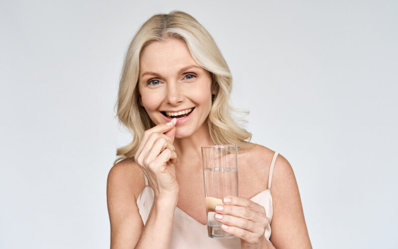 Happy middle aged 50s woman holding pill and glass of water taking dietary supplements