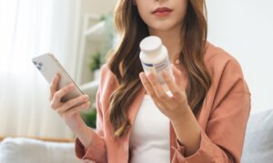 Health care asian young woman using smart phone for reading, searching prescription on bottle medicine, pill label text about information online, instructions side effects, pharmacy medicament concept