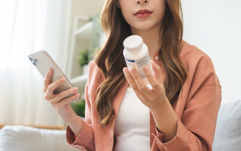 Health care asian young woman using smart phone for reading, searching prescription on bottle medicine, pill label text about information online, instructions side effects, pharmacy medicament concept