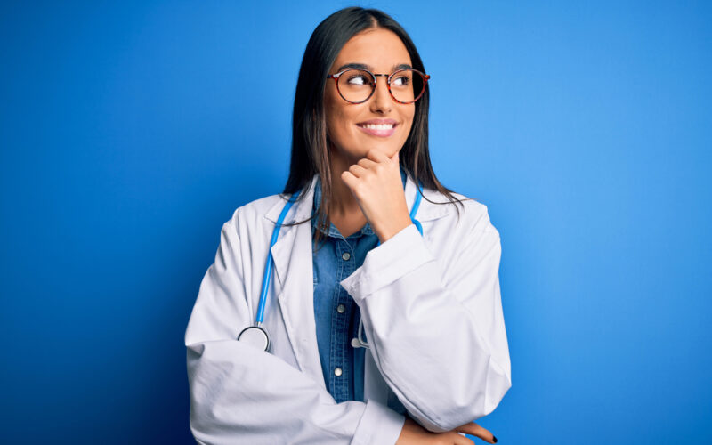 Young beautiful doctor woman wearing stethoscope and glasses over blue background with hand on chin thinking about question, pensive expression. Smiling with thoughtful face. Doubt concept.