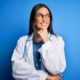 Young beautiful doctor woman wearing stethoscope and glasses over blue background with hand on chin thinking about question, pensive expression. Smiling with thoughtful face. Doubt concept.