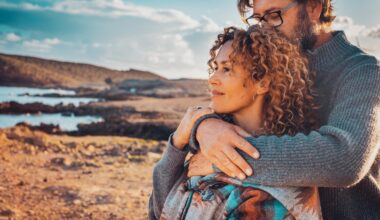 A couple embraces outdoors by the ocean at sunset, with the woman looking ahead thoughtfully while the man gently holds her from behind, symbolizing intimacy, connection, and comfort.
