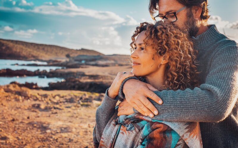 A couple embraces outdoors by the ocean at sunset, with the woman looking ahead thoughtfully while the man gently holds her from behind, symbolizing intimacy, connection, and comfort.
